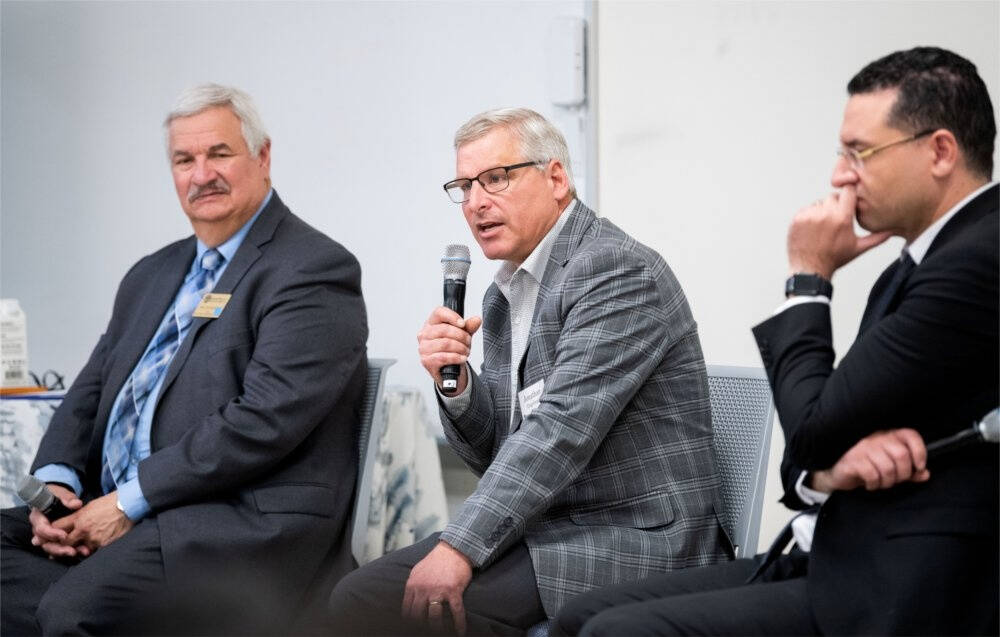 Three men hold microphones wearing official attires seem to address people in a room.
